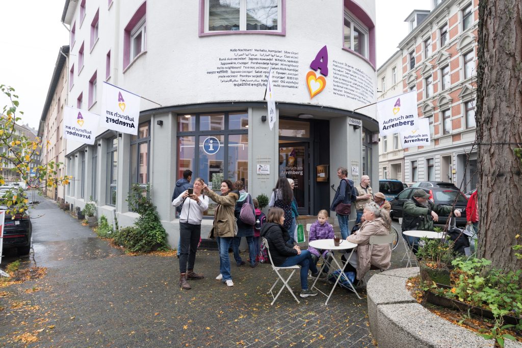 Menschen stehen und sitzen vor dem Vereinsgebäude des Aufbruch am Arrenberg e.V.; auf der Fassadse sieht man das Vereinslogo, das eine Rakete in Form eines lilafarbenen A darstellt, der Feuerschweif besteht aus konzentrischen Herzen in weiß, gelb und orange. Neben dem Logo ist die Begrüßung »Hallo Nachbars« in 50 Sprachen angebracht.