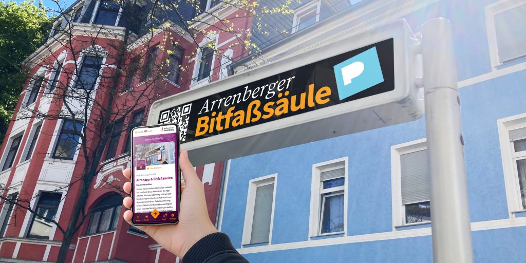 Ein Straßendisplay vor einer Arrenberger Häuserfassade zeigt einen Beitrag mit dem Titel »Bitfaßsäule«; im Vordergrund hält eine Hand ein Mobiltelefon, auf dem der gleiche Beitrag in der App gezeigt wird.