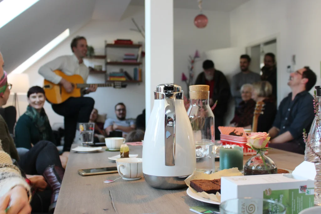Ein gedeckter Kaffeetisch einer Privatwohnung, im Hintergrund unerhalten sich Leute, einer von ihnen spielt Gitarre.
